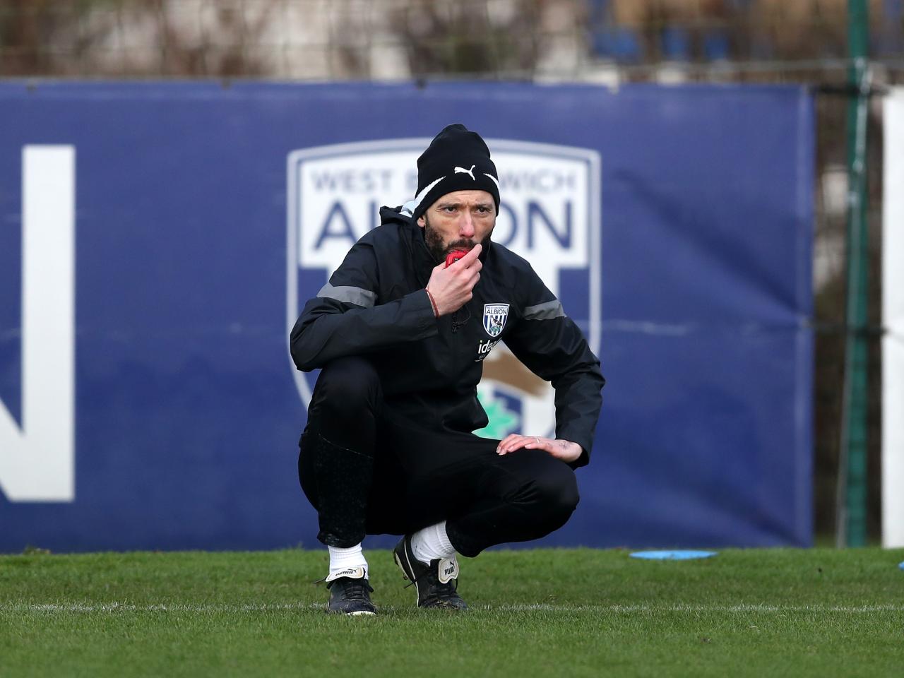 Carlos Corberán crouched down watching training in front of an Albion badge in the background