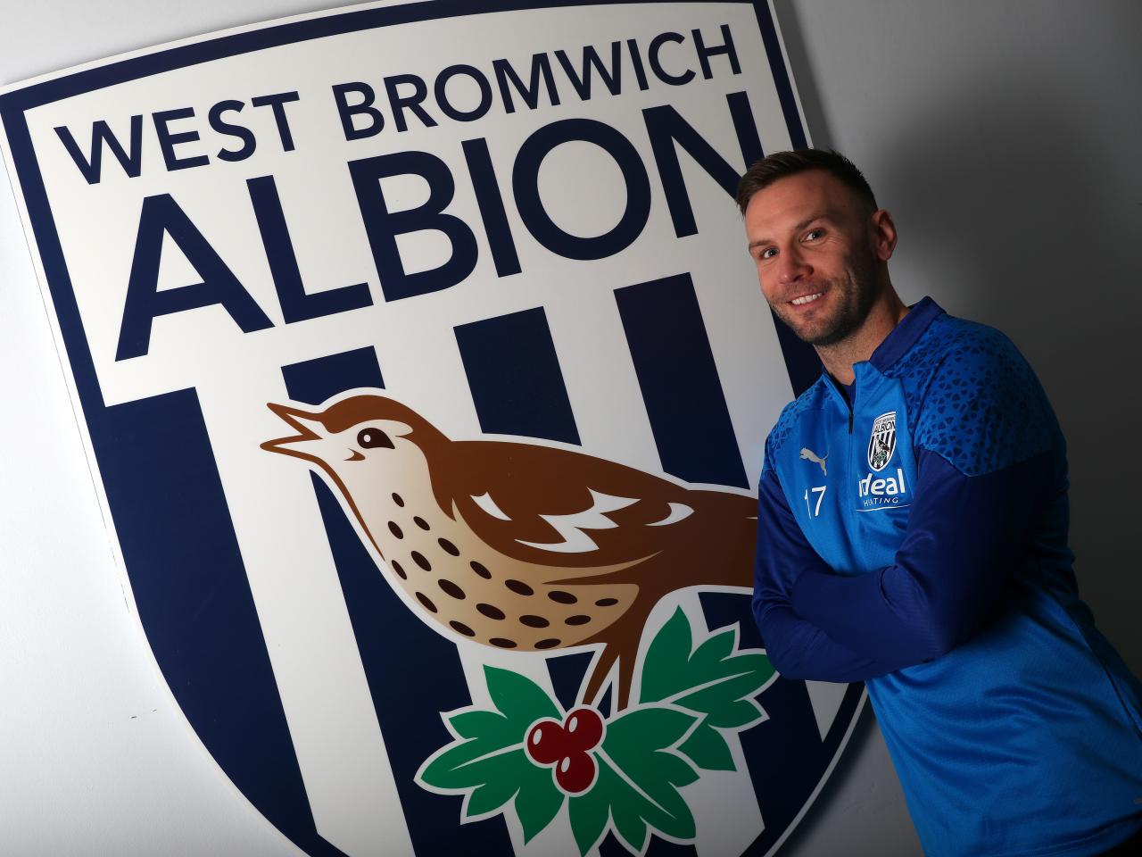 Andi Weimann smiling at the camera stood to the right of a big WBA badge on the wall