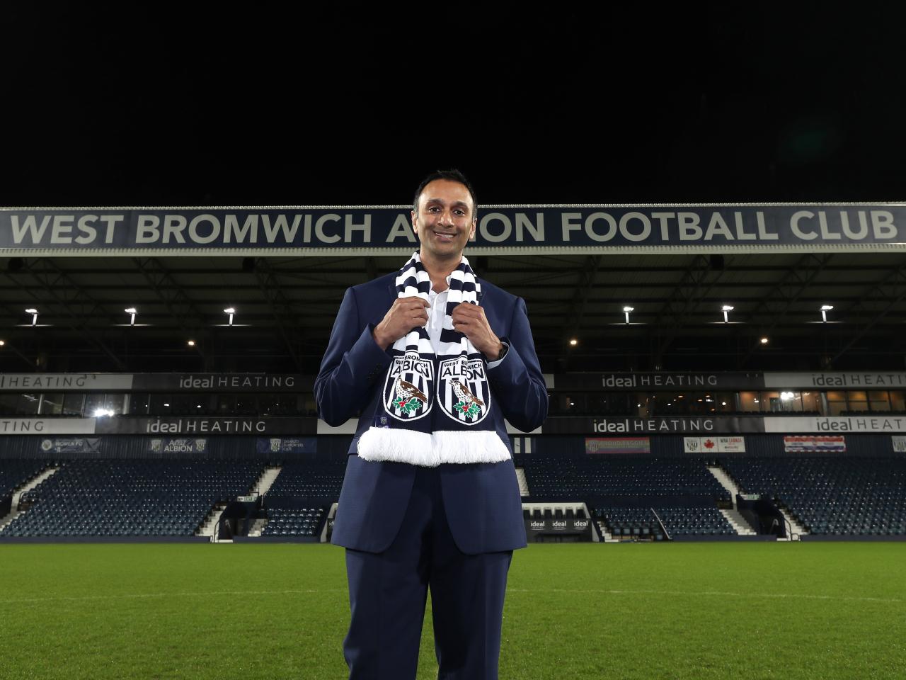 Shilen Patel stood on the pitch at The Hawthorns with the West Stand behind him and an Albion scarf wrapped around his neck