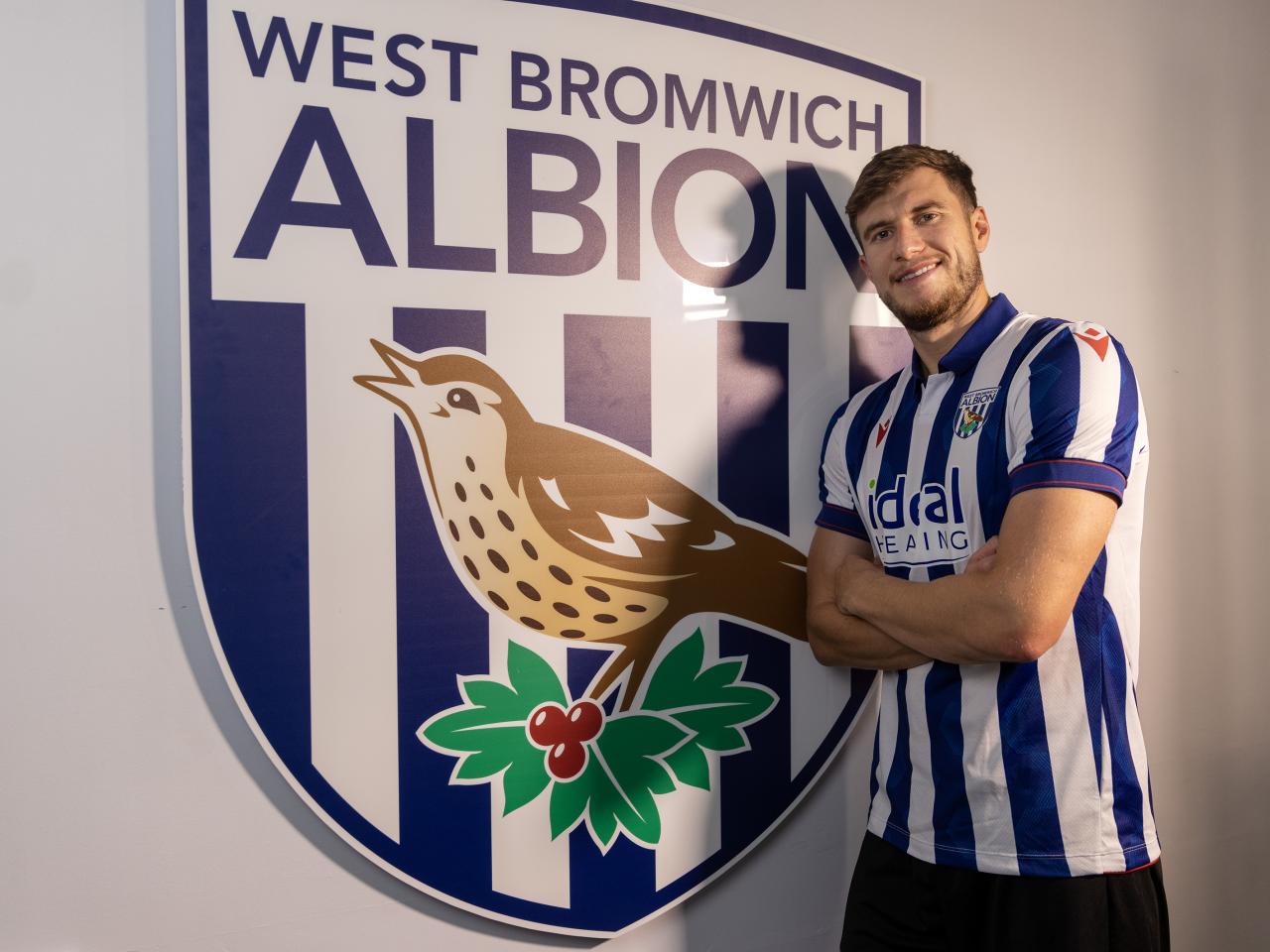 Paddy McNair smiling at the camera while wearing a home shirt stood in front of an Albion badge