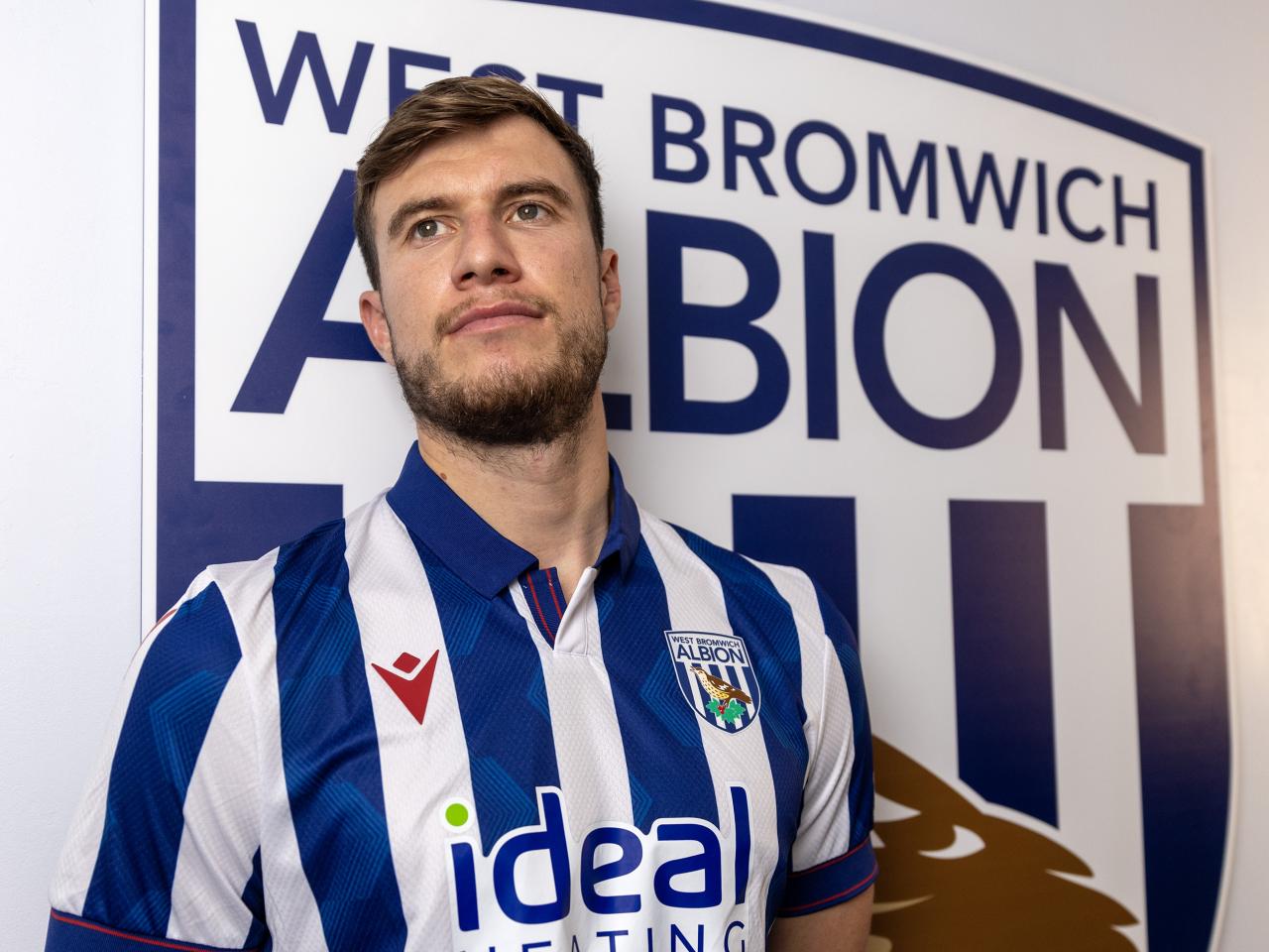 Paddy McNair looking away from the camera while wearing a home shirt stood in front of an Albion badge