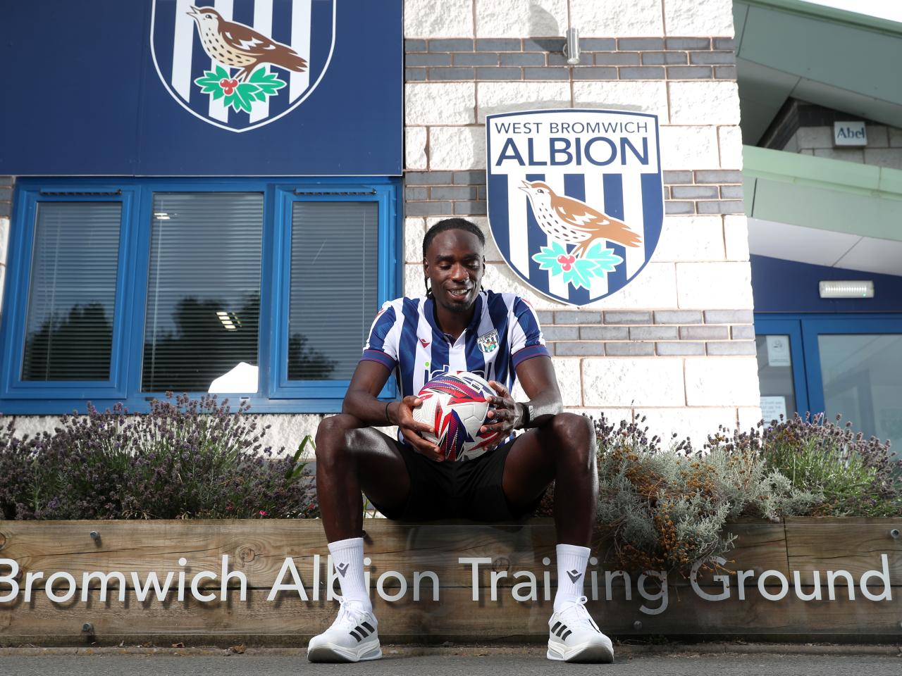 Devante Cole smiling while looking down at a ball sat outside the training ground entrance in a home shirt 