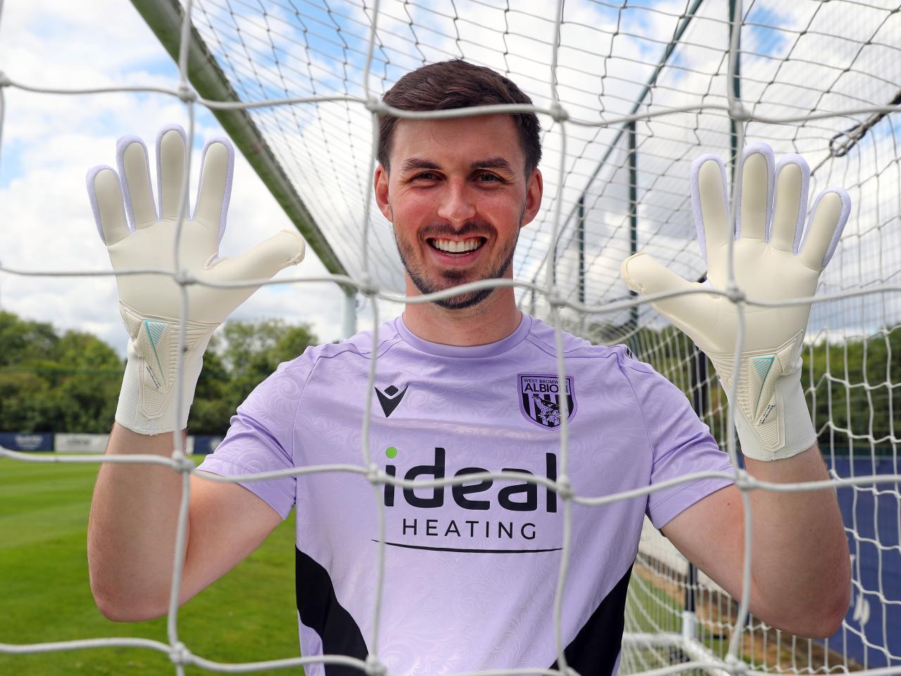 Joe Wildsmith smiling at the camera while holding his gloves up and looking through a goal net
