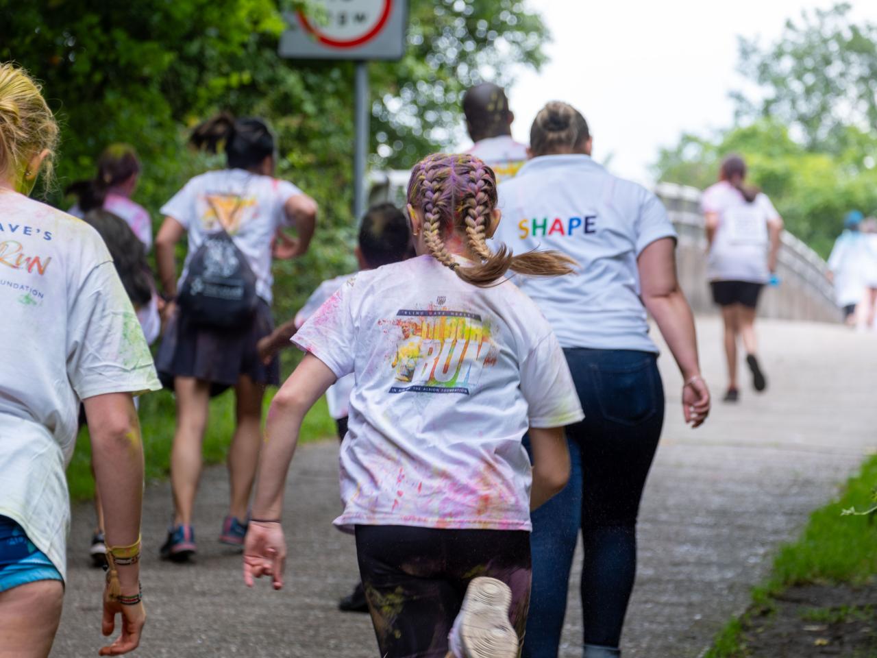 Back of Runner heading town motorway bridge, with Colour Run branding on display & SHAPE Festival displayed on one person back.