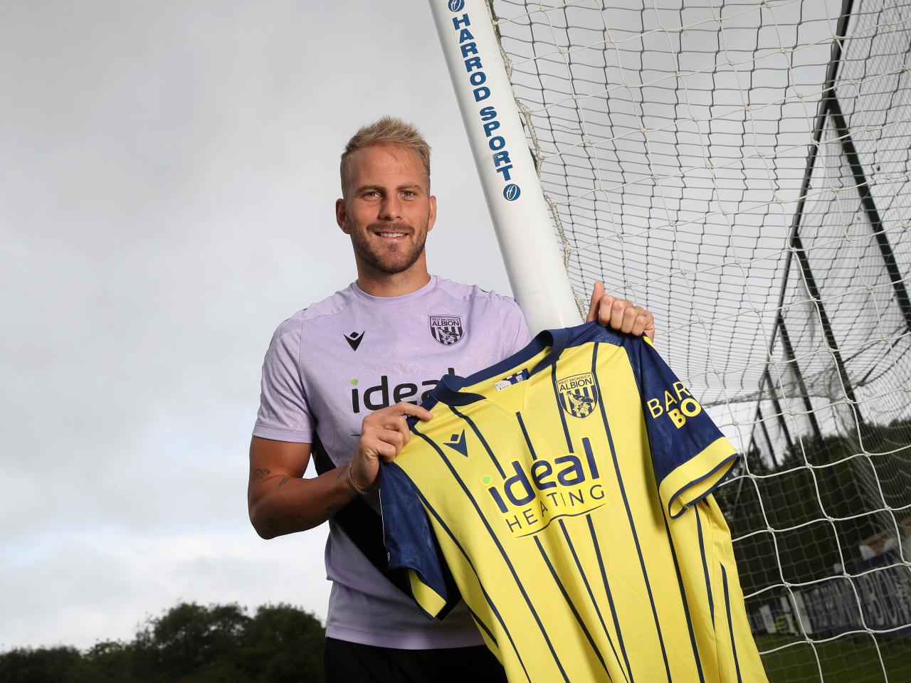 Uroš Račić smiling at the camera while stood holding up a yellow away shirt 