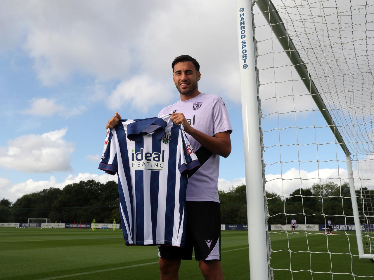 Gianluca Frabotta smiling at the camera while stood against a goal post holding up a home shirt 