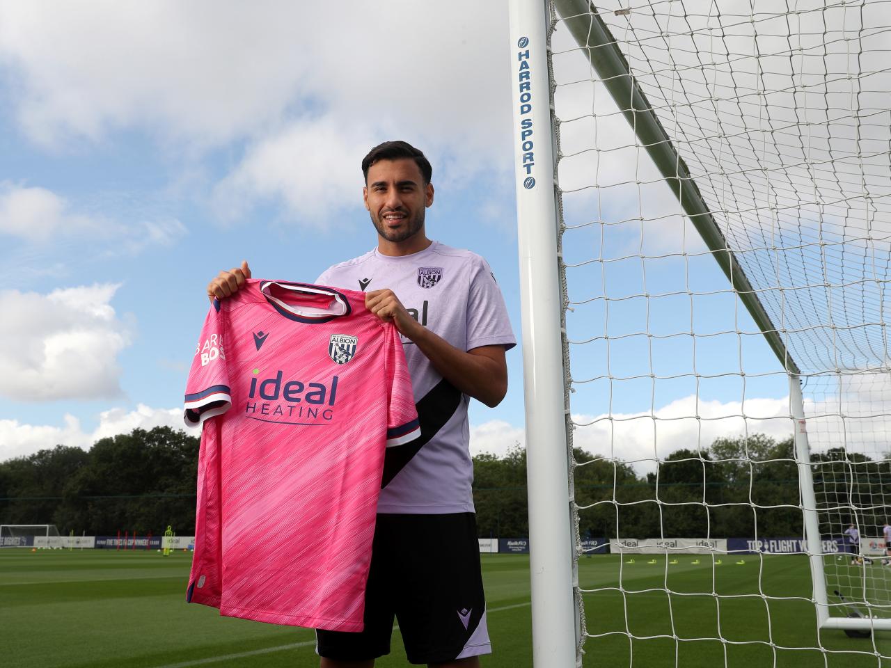 Gianluca Frabotta smiling at the camera while stood against a goal post holding up a pink away shirt