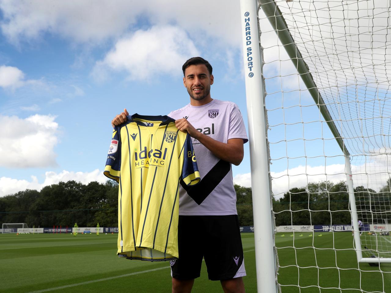 Gianluca Frabotta smiling at the camera while stood against a goal post holding up a yellow away shirt
