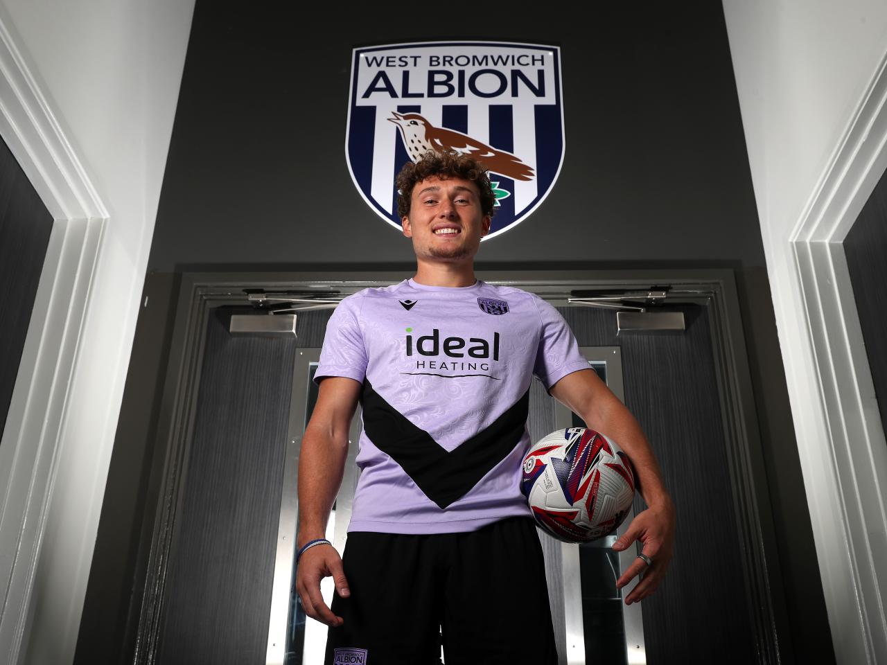 Callum Styles smiling at the camera while holding a ball while stood in a corridor 