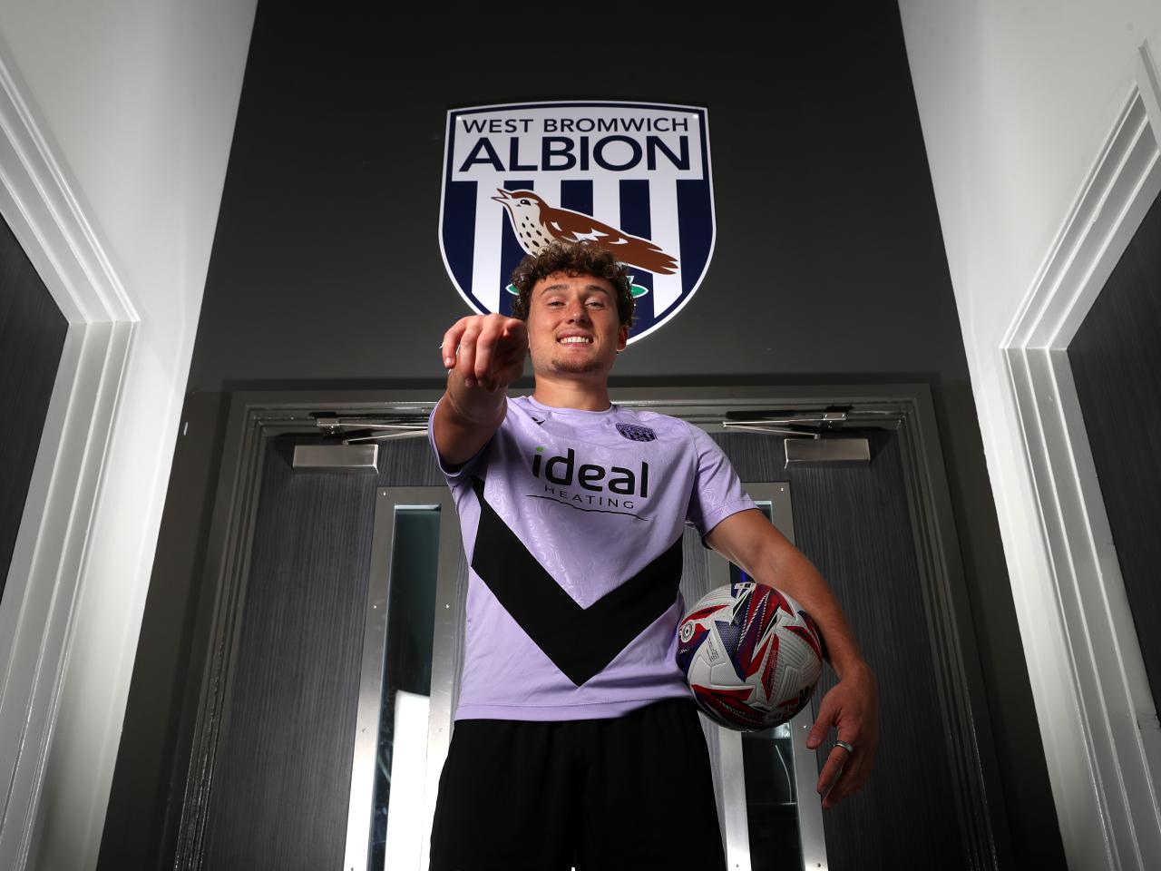 Callum Styles smiling at the camera while holding a ball and pointing at the camera in a corridor 