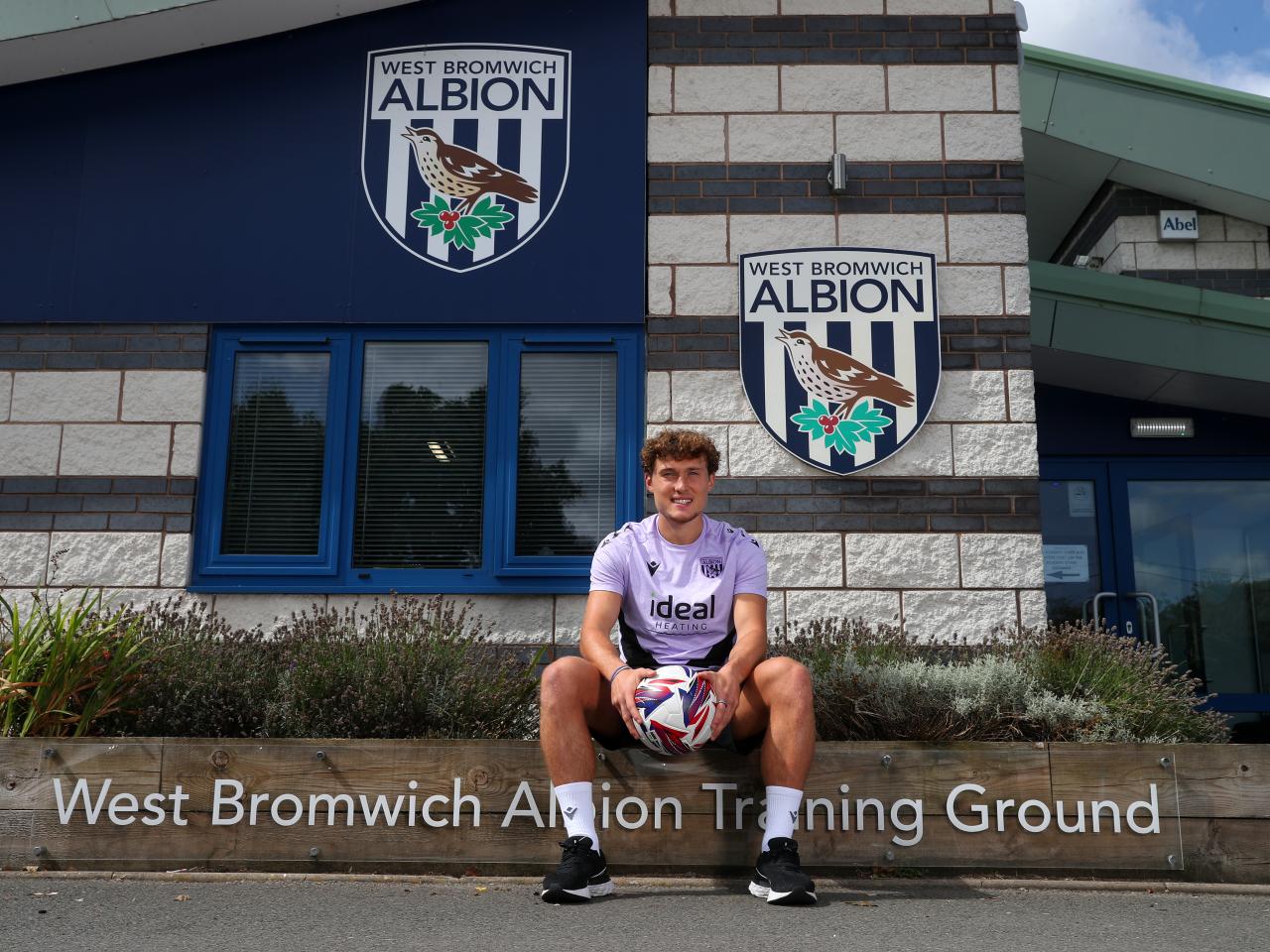 Callum Styles smiling at the camera while holding a ball sat outside the front of the training ground 