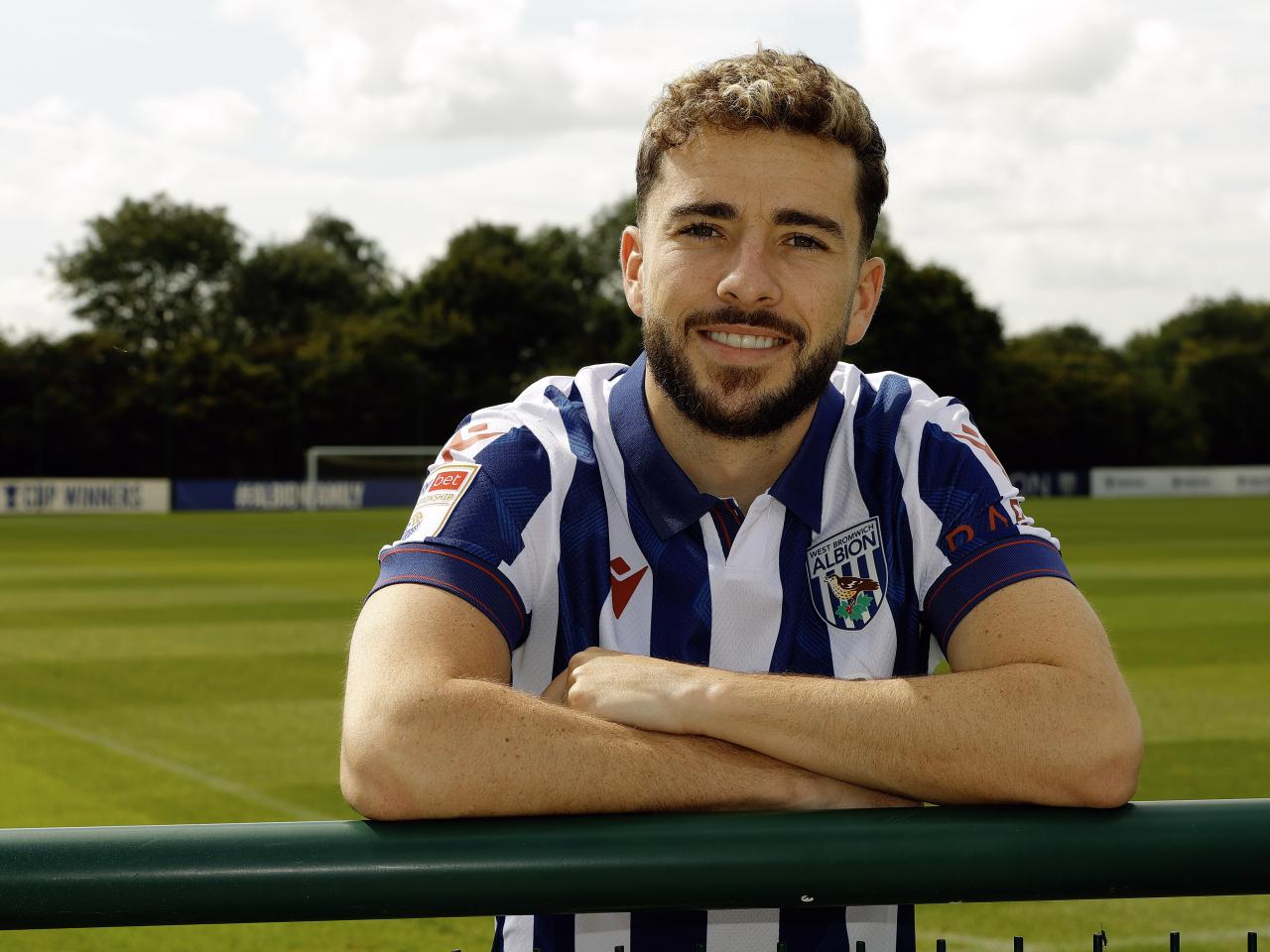 Mikey Johnston leaning over a barrier with his arms folded smiling at the camera while wearing a home shirt 