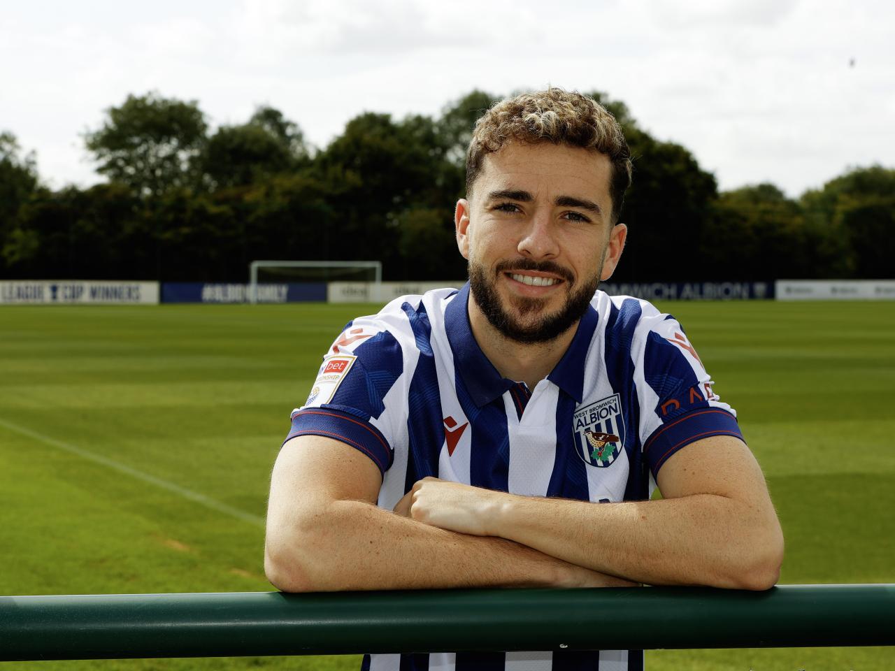 Mikey Johnston leaning over a barrier with his arms folded smiling at the camera while wearing a home shirt 