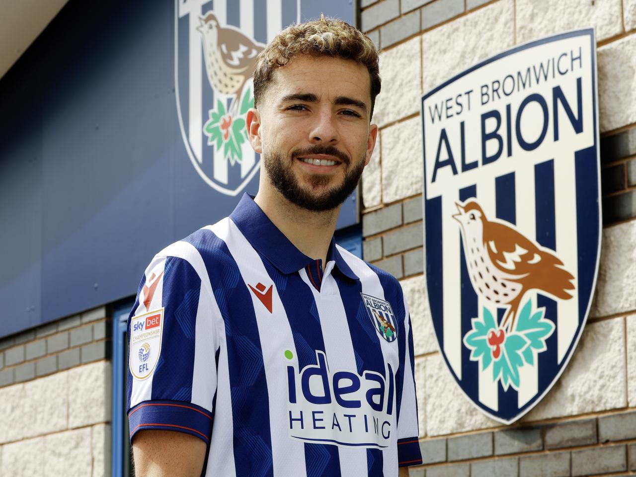 Mikey Johnston smiling at the camera while stood outside the training ground in a home shirt 