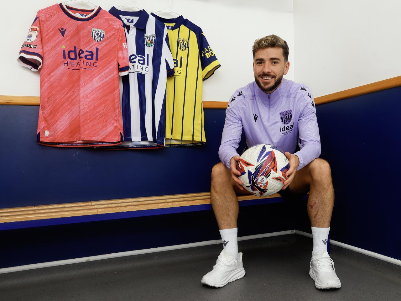 Mikey Johnston sat in a dressing room holding a ball and smiling with all three shirts hanging up behind him