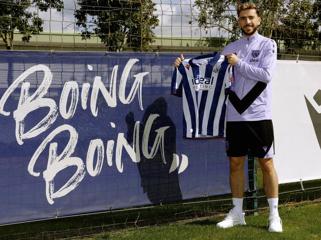 Mikey Johnston smiling at the camera holding up a home shirt stood next to a sign which says boing boing 