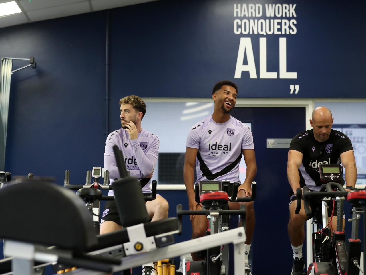 Mason Holgate on a bike in the gym sat next to Mikey Johnston and a member of staff with a smile on his face