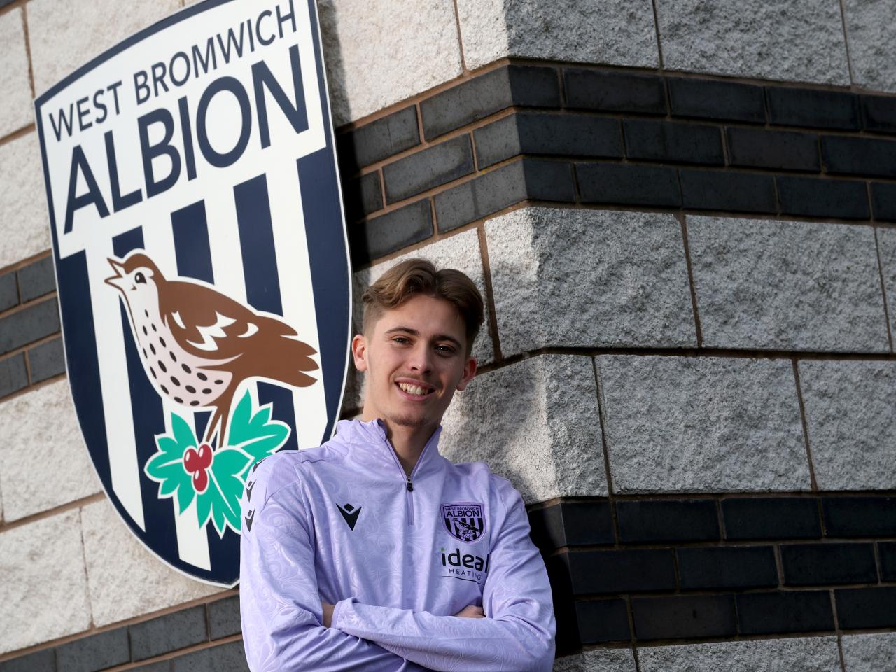Isaac Price stood up against a wall in Albion training gear next to a WBA badge