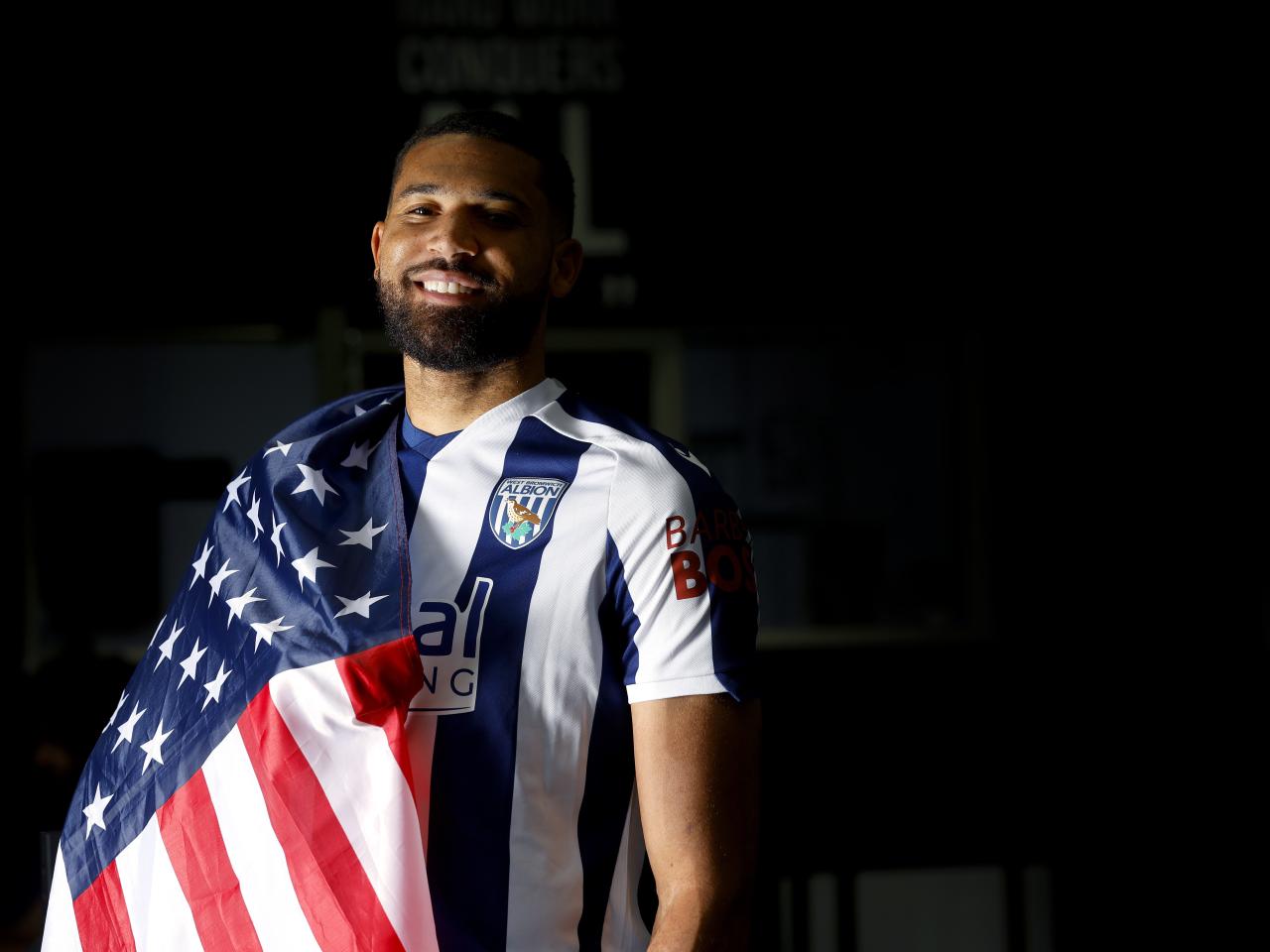 George Campbell smiling at the camera while in a home WBA shirt with a USA flag wrapped around his shoulder