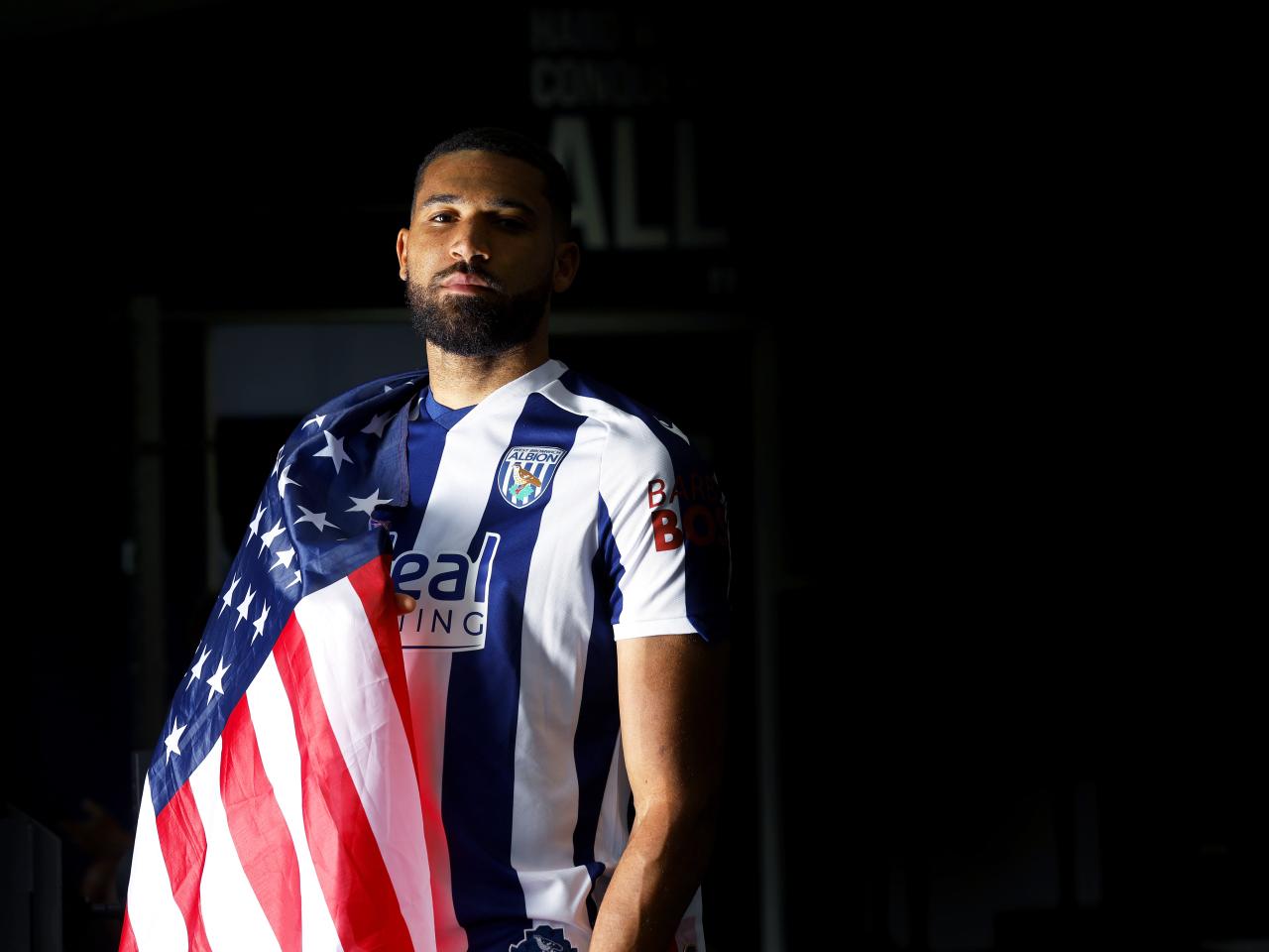 George Campbell looking at the camera while in a home WBA shirt with a USA flag wrapped around his shoulder