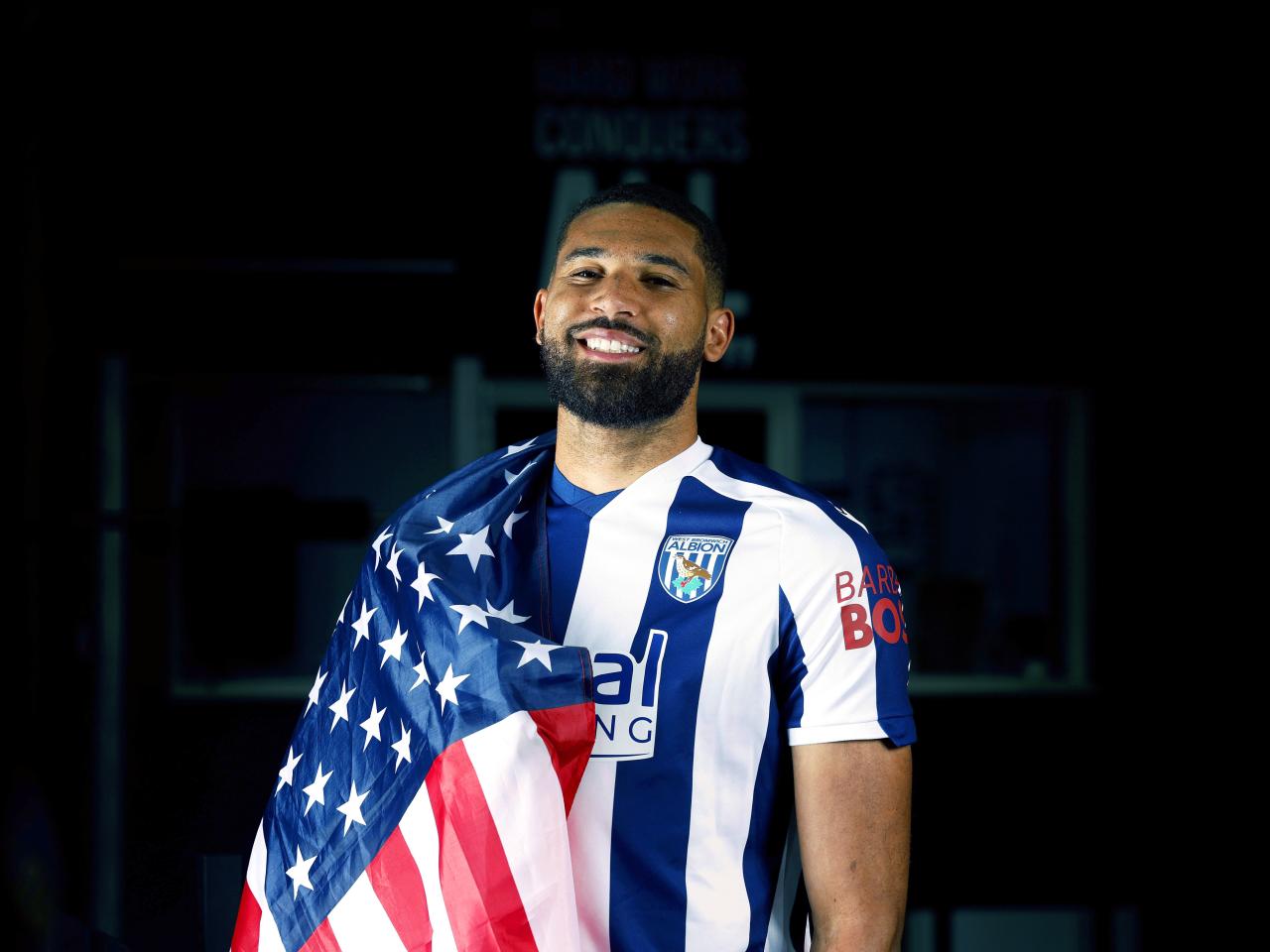 George Campbell smiling at the camera while in a home WBA shirt with a USA flag wrapped around his shoulder