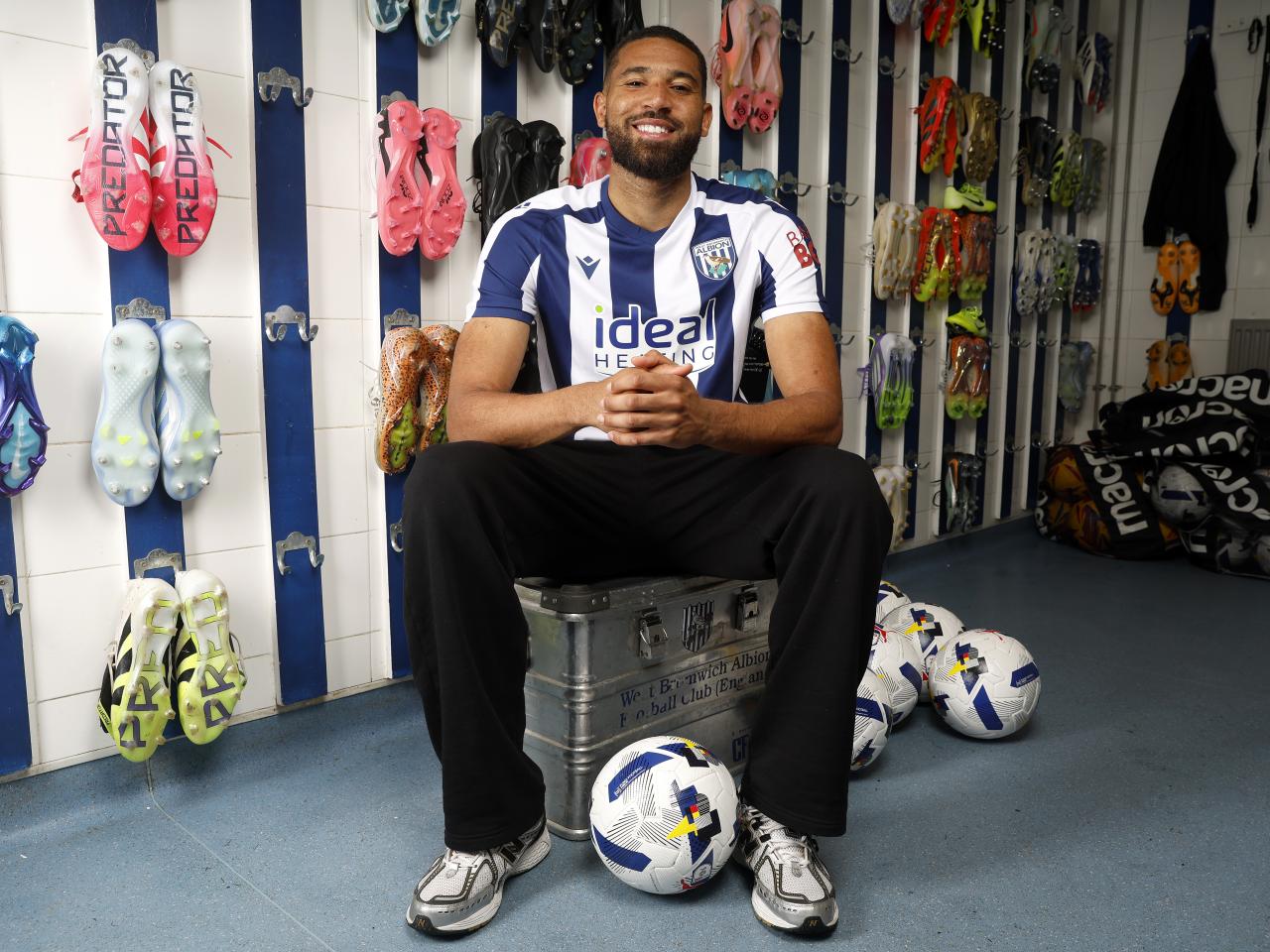 George Campbell sat in the boot room in a home WBA shirt smiling at the camera with footballs by his feet 