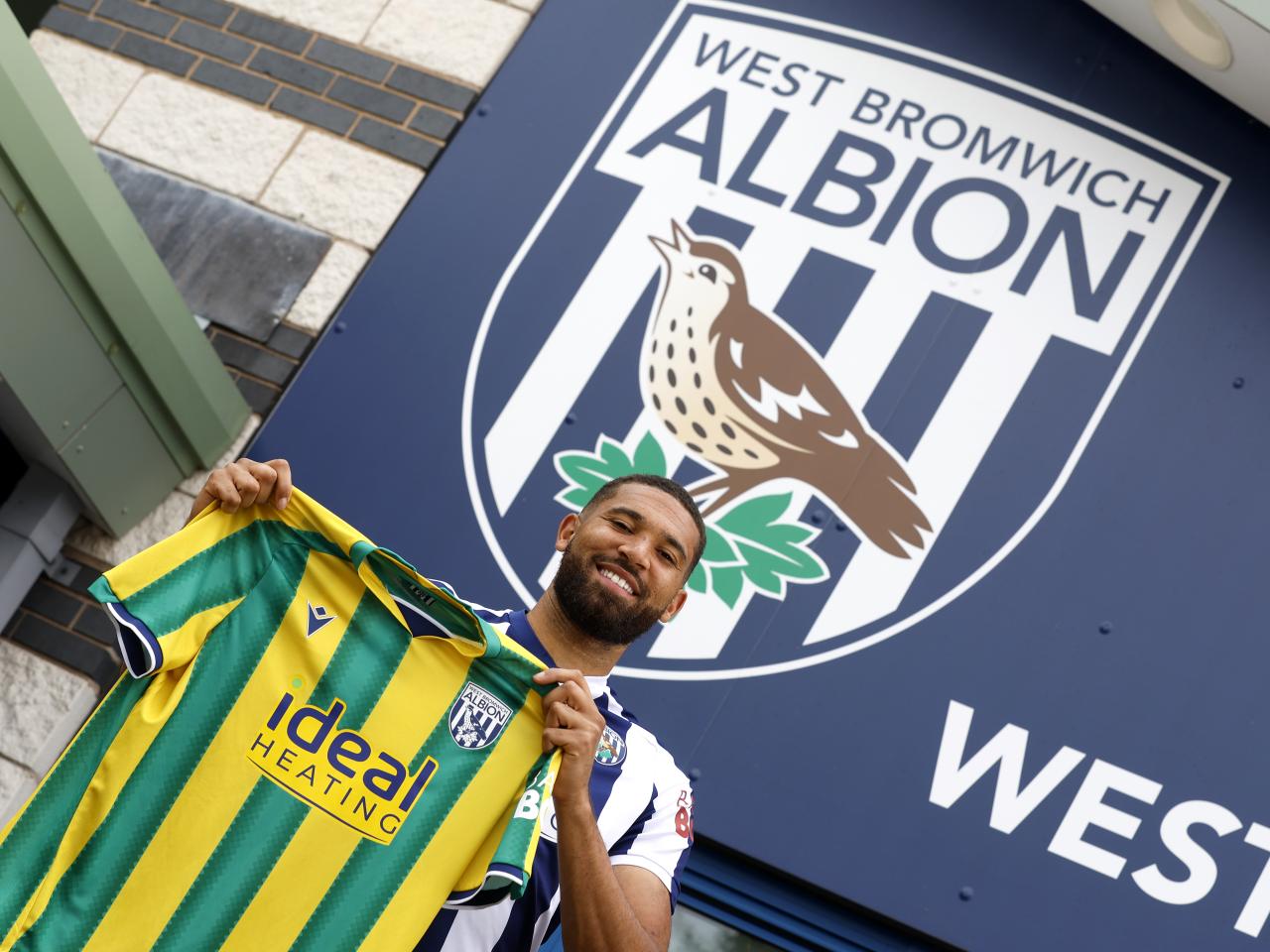 George Campbell holding up an away WBA shirt and smiling at the camera while stood in front of WBA branding