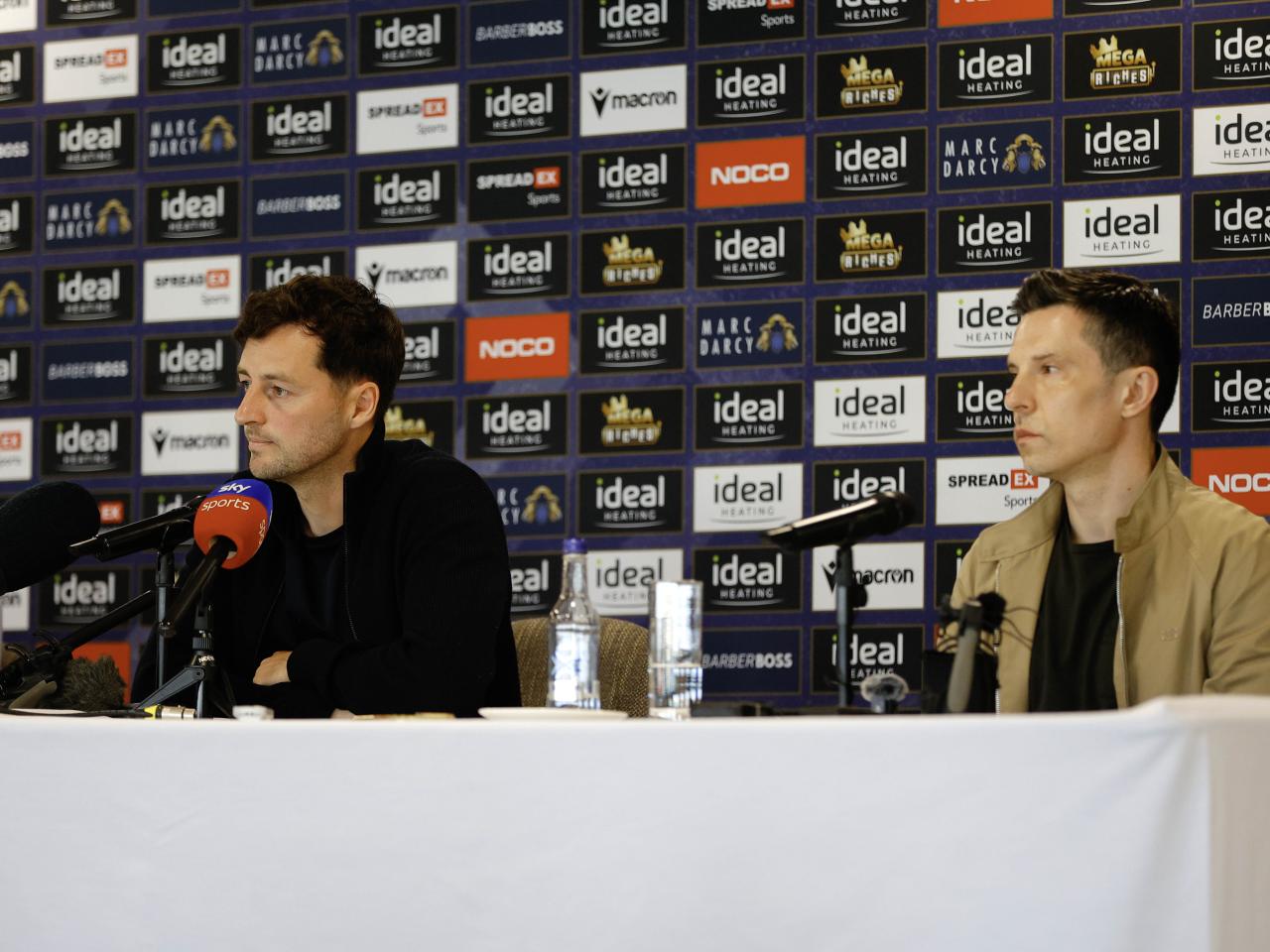 Ryan Mason and Andrew Nestor talking to members of the media in front of a press conference sponsor board and behind a number of microphones