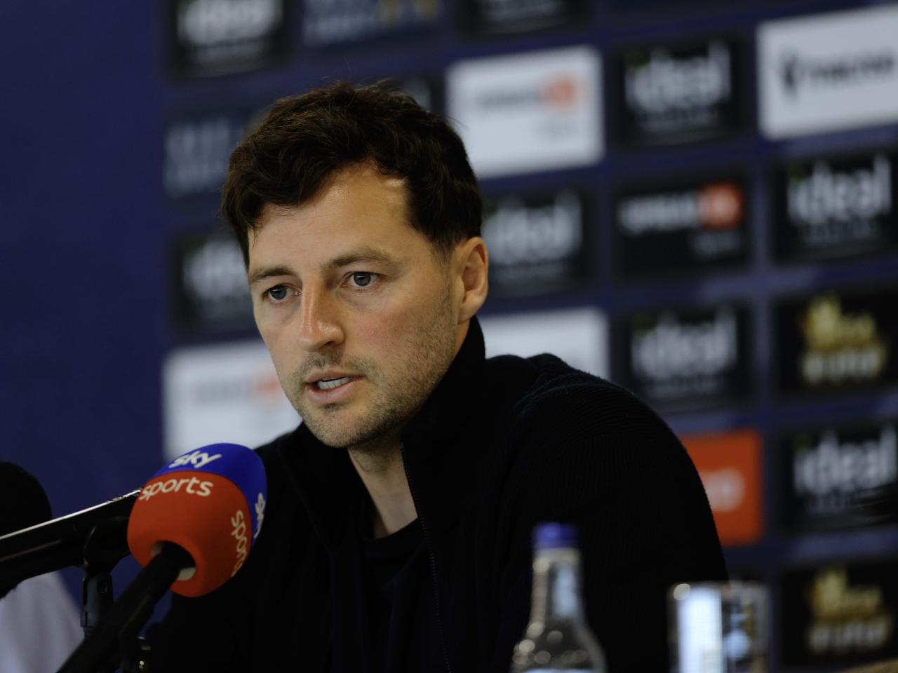 Ryan Mason talking to members of the media in front of a press conference sponsor board and behind a number of microphones