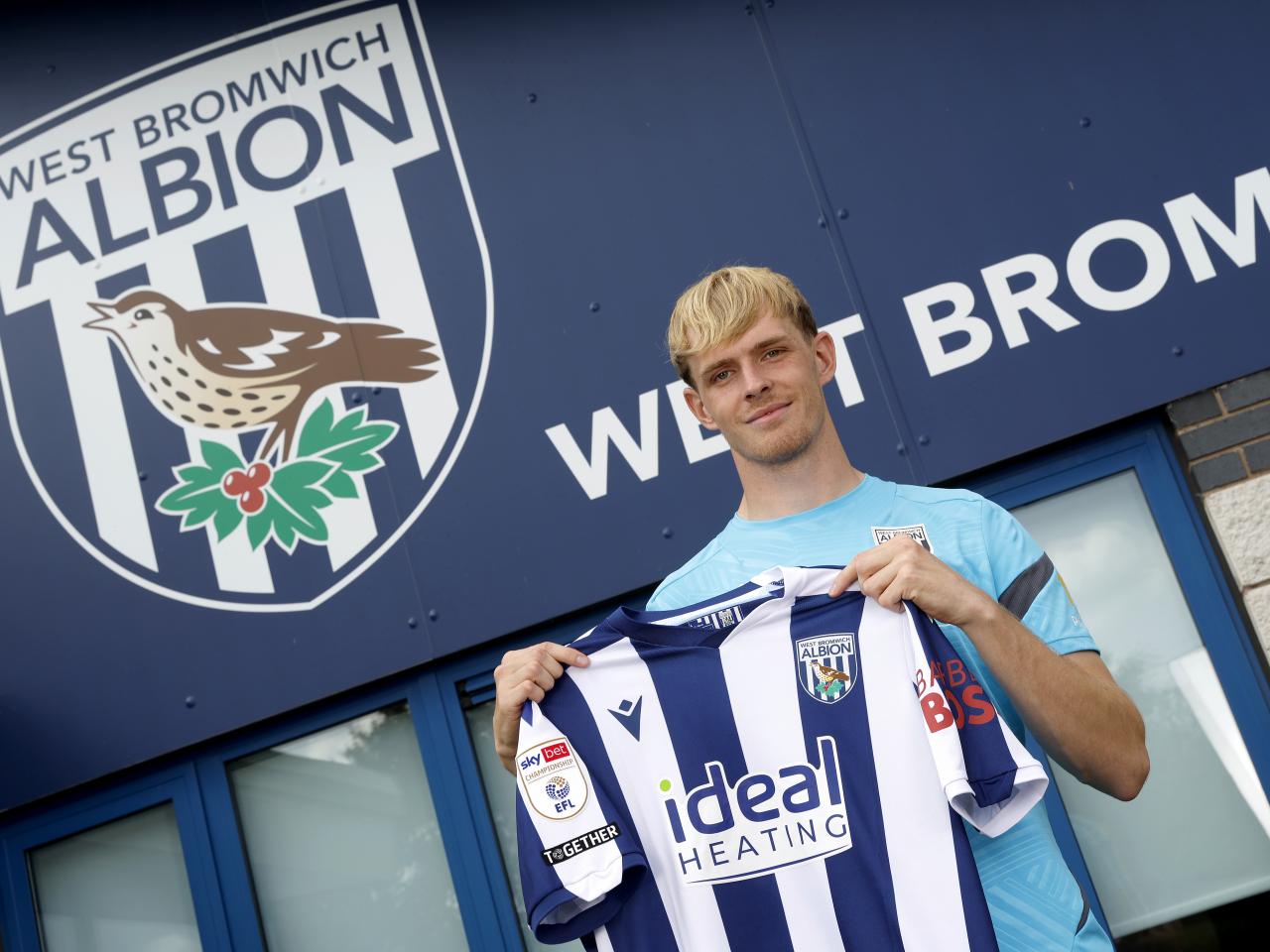 Toby Collyer smiling and looking at the camera while stood outside the training ground holding up a WBA shirt 