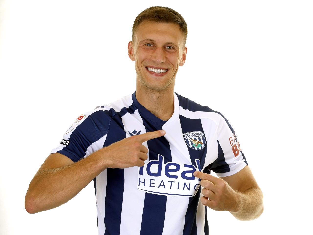 Krystian Bielik smiling at the camera and pointing to the badge on the home shirt 