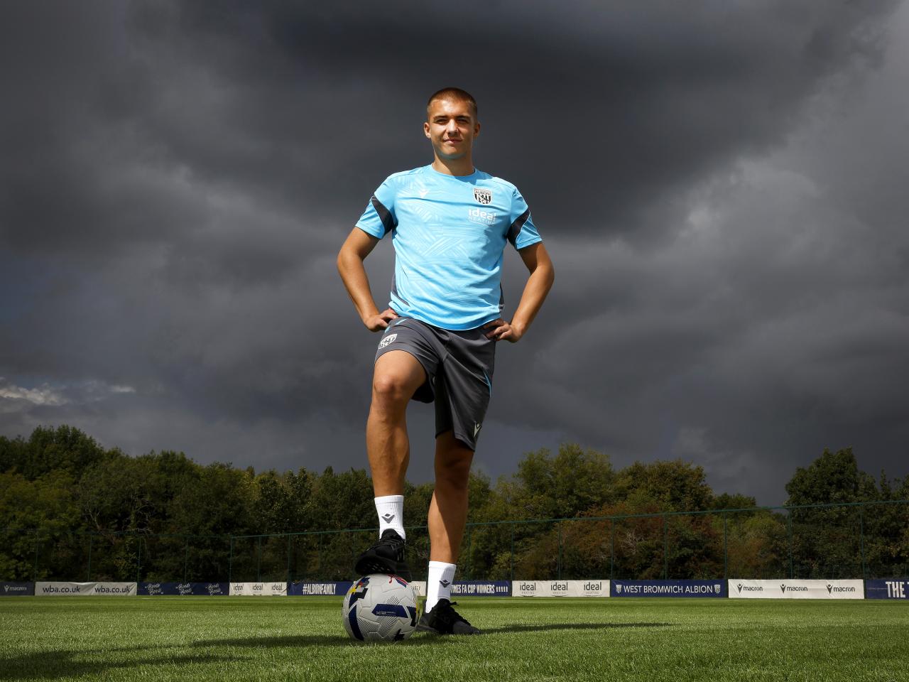 Alfie Gilchrist stood with his foot on a ball and his hands on his hips out on a training pitch smiling at the camera 