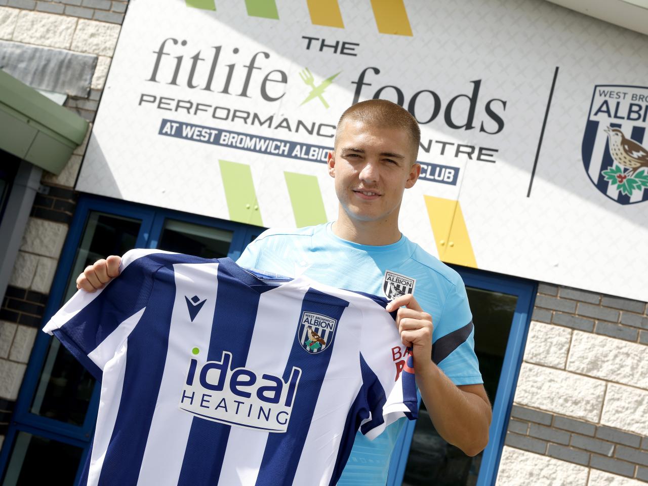 Alfie Gilchrist outside the front of the training ground holding up a home shirt smiling at the camera 