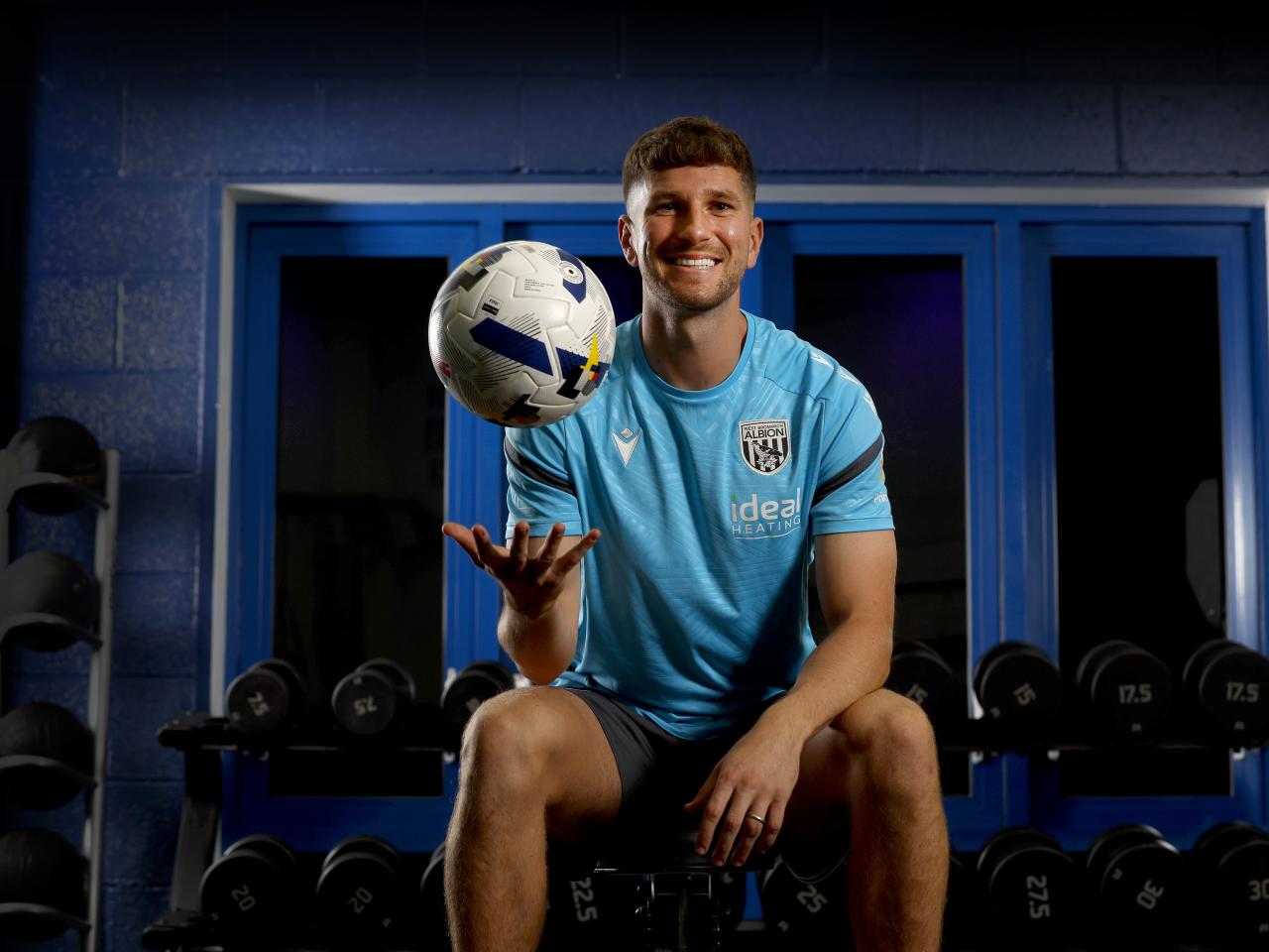 Chris Mepham smiling at the camera while holding a ball and being sat down in the gym