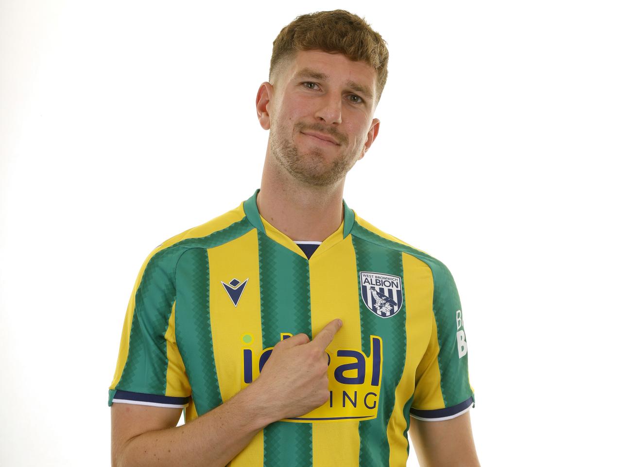 Chris Mepham in the green and yellow away kit smiling at the camera and pointing at the badge