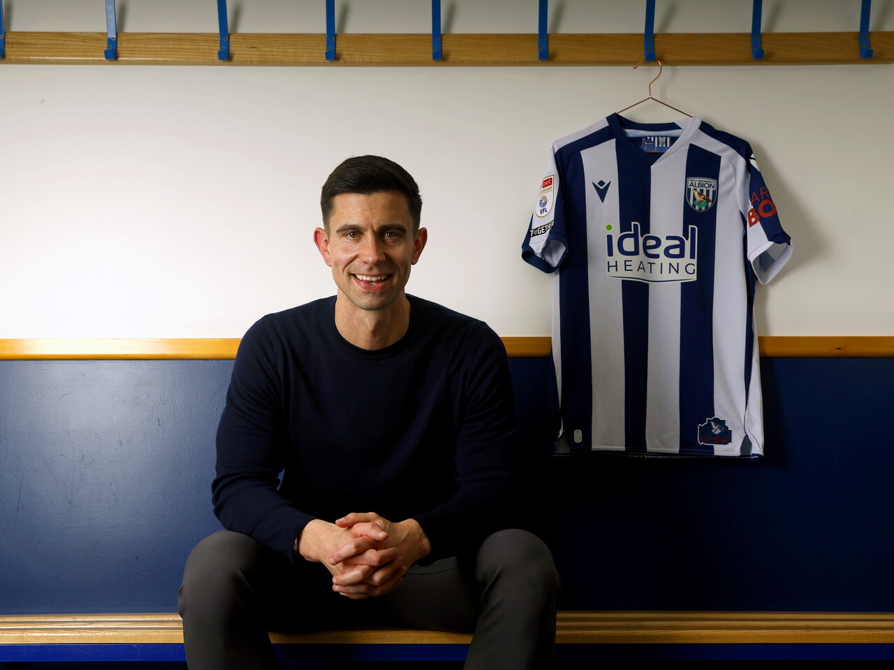 Eric Ramsay smiling at the camera while sat down in a dressing room with a home shirt hanging next to him