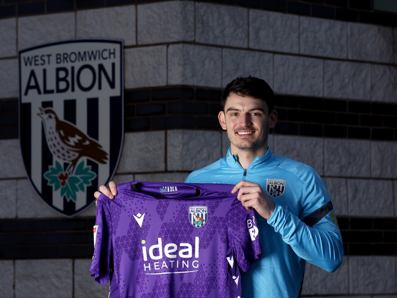 Max O'Leary smiling at the camera while holding up a WBA shirt 