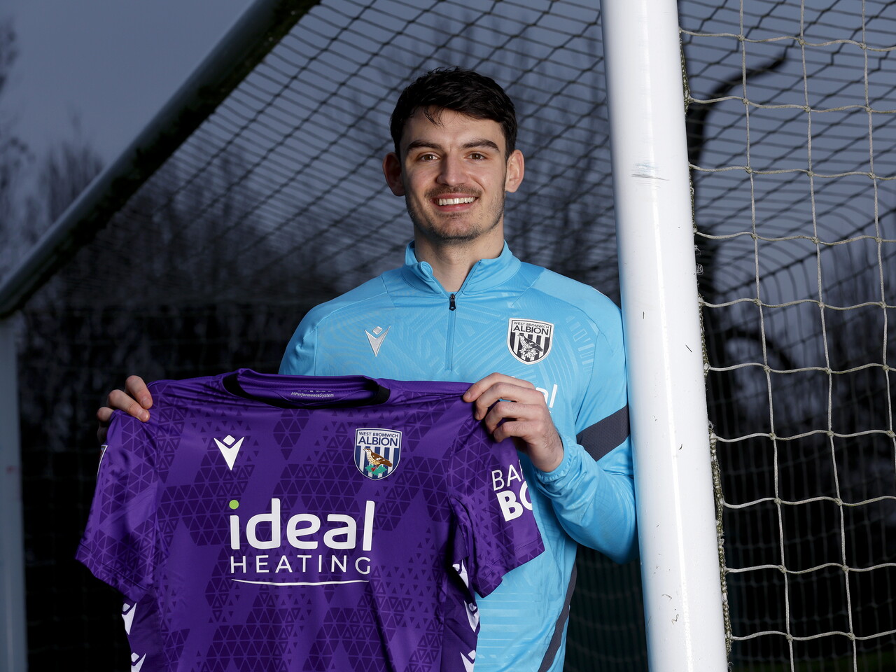 Max O'Leary smiling at the camera while leaning against a goal post and holding up a WBA shirt 
