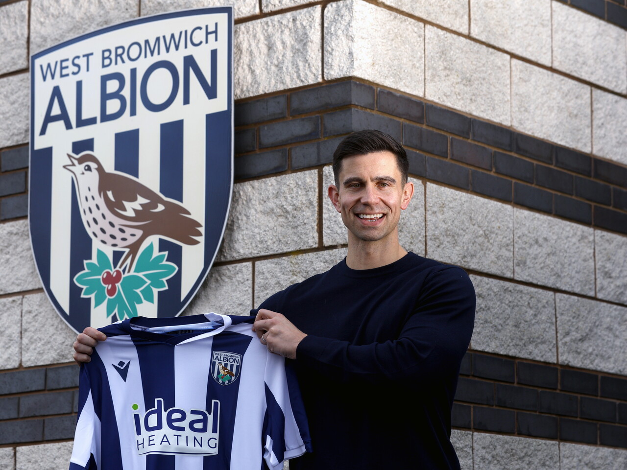 Eric Ramsay smiling at the camera while stood next to a WBA badge and holding a home WBA shirt