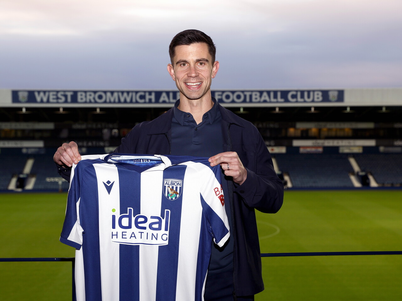 Eric Ramsay smiling at the camera and holding up a home WBA shirt at The Hawthorns