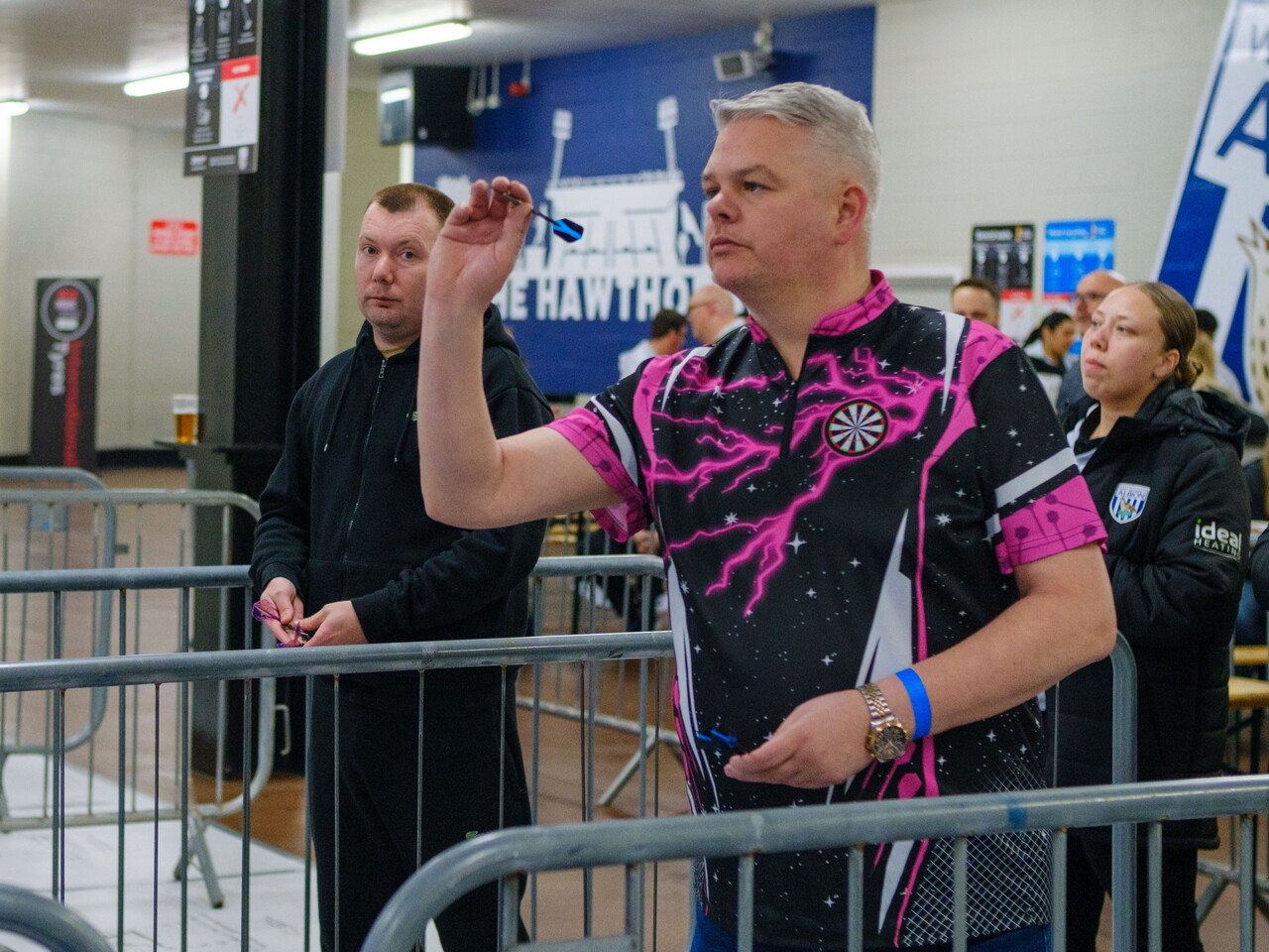 Adult darts player taking aim with another player looks on.