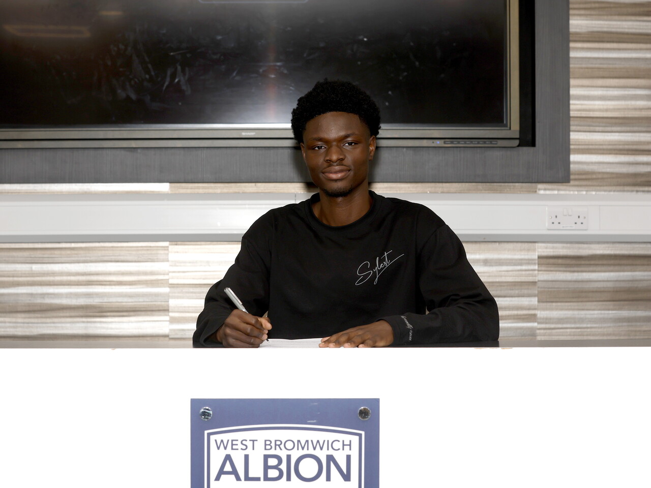 Abdul Abudu smiling at the camera while signing a professional contract