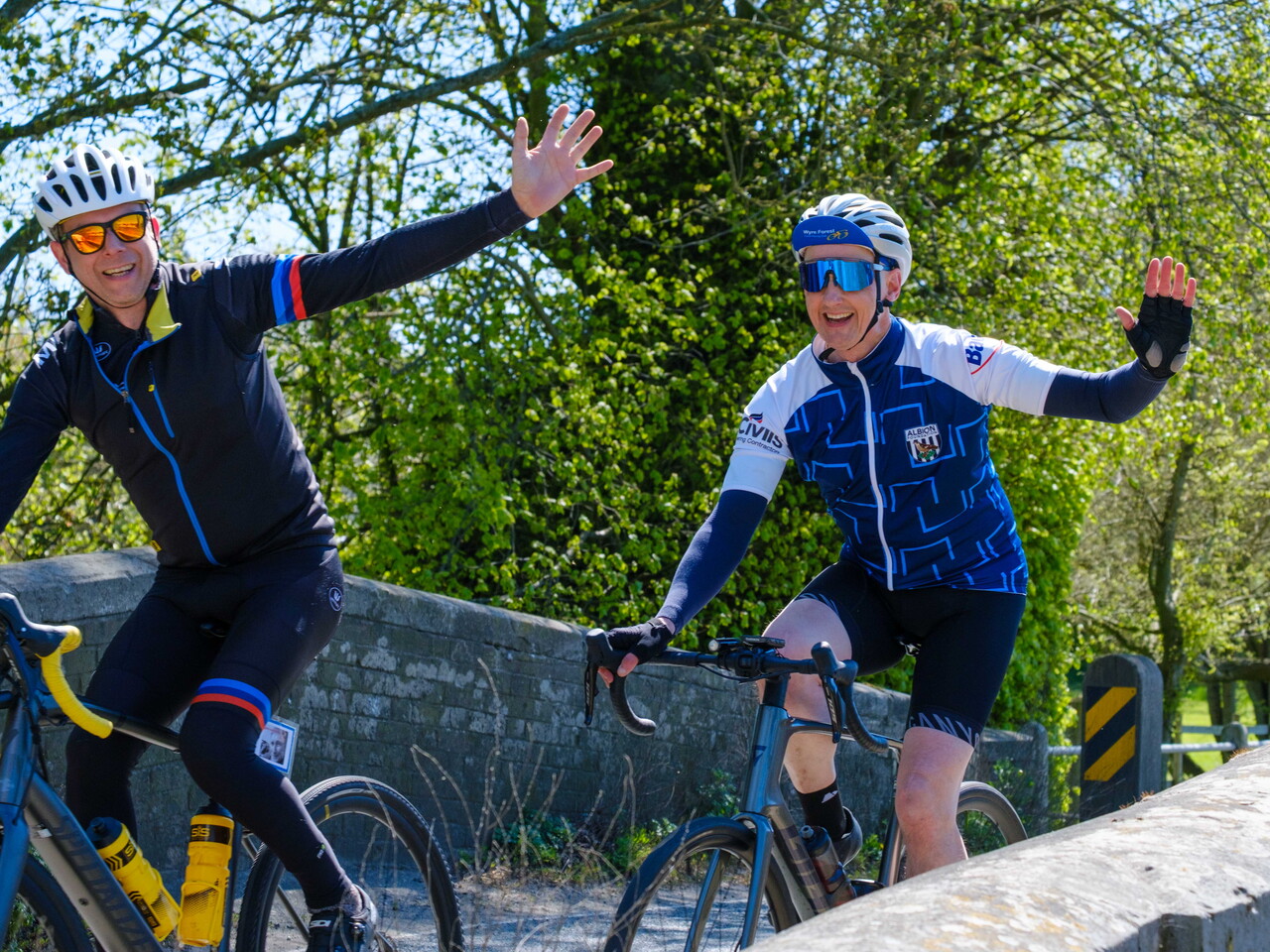 Cyclists waving as they cross a small bridge.