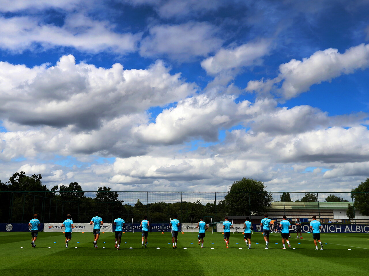 A general shot of several players running in a straight line at the training ground with blue and cloudy skies above