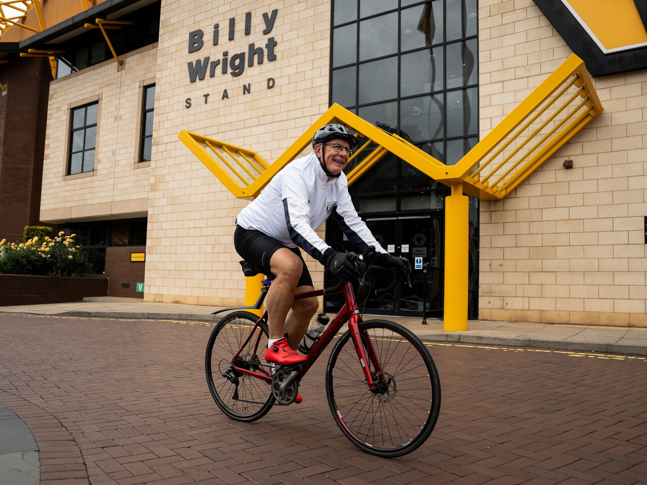 A cyclist wearing a white jersey, black shorts, and a helmet rides a red road bike outside the Billy Wright Stand entrance at a football stadium, with yellow architectural features visible behind him.
