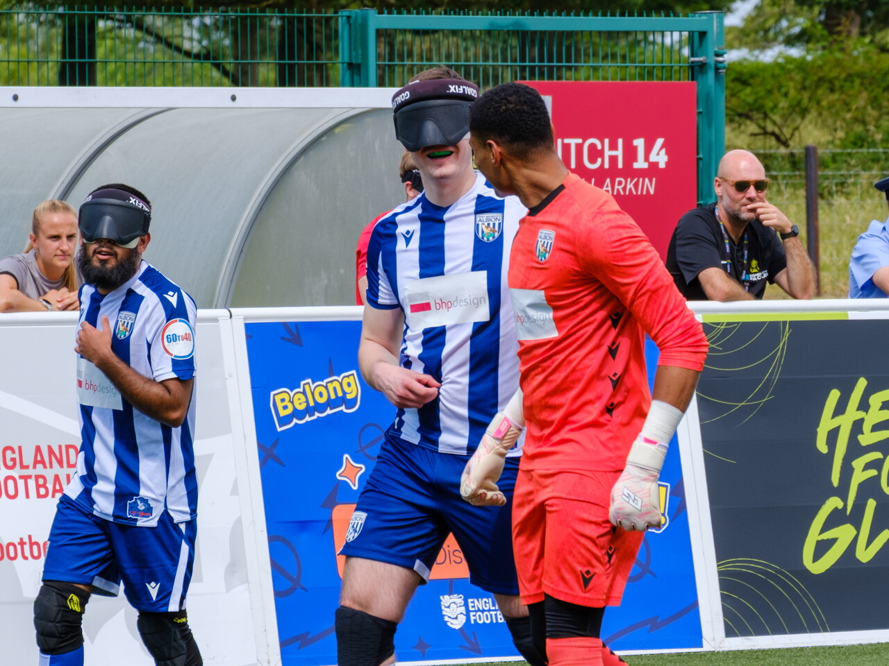 Efe Shimwell, Eesa Amjid and Owen Locke  gather near the sideline—two in blue-and-white striped kits wearing eye masks and one in a bright red goalkeeper kit—standing beside advertising boards and a team bench, with spectators and staff visible behind a pitch board.