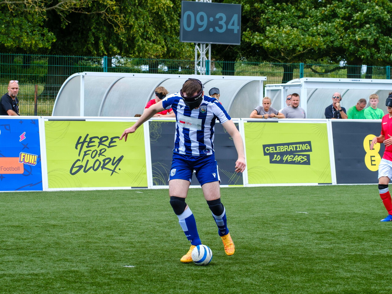 Efe Shimwell in a blue-and-white striped West Bromwich Albion kit dribbles a ball on an outdoor pitch while wearing an eye mask. A scoreboard behind shows “09:34,” and advertising boards display text including “Here for Glory” and “Celebrating 10 Years,” with spectators watching from behind the barrier.