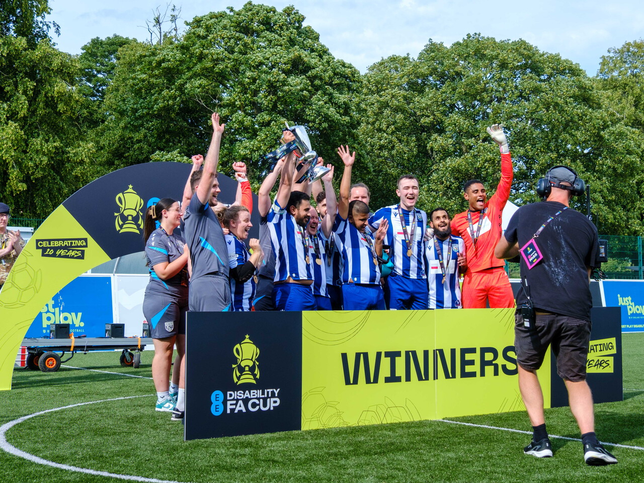 WBA Blind players on outdoor pitch, raising a trophy and their arms in celebration. A large sign in front reads “Winners” alongside the EE Disability FA Cup logo, while a camera operator films the moment and trees fill the background.