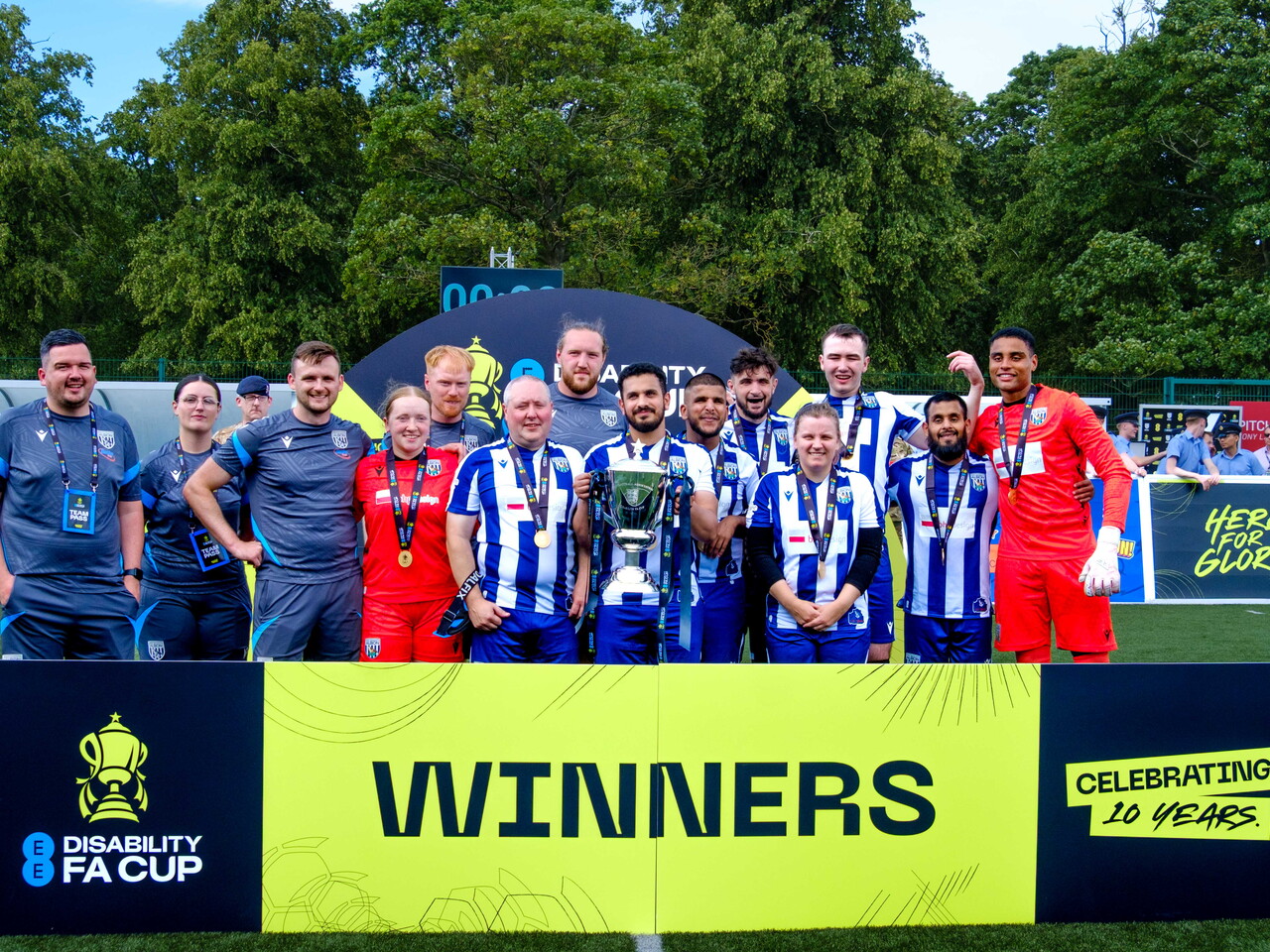 WBA Blind players on an outdoor pitch, Captain Mo Ahmed holding the trophy. A large sign in front reads “Winners” alongside the EE Disability FA Cup logo, and trees fill the background.