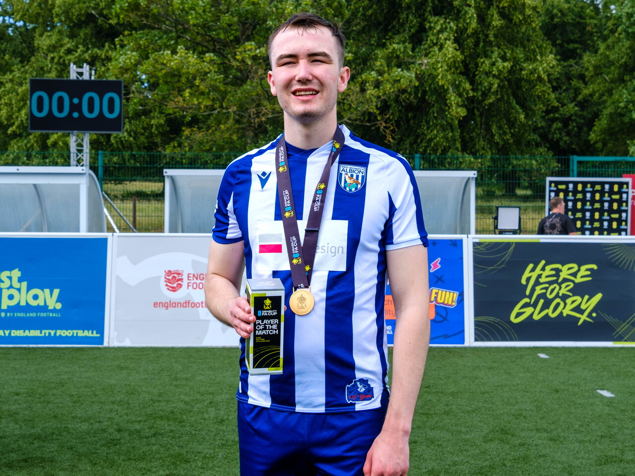 Efe Shimwell  stands on an outdoor football pitch wearing a blue-and-white striped kit, holding a Player of the Match award and wearing a medal on a ribbon around the neck. Advertising boards and a small stand are visible behind, with trees and a fence in the background.